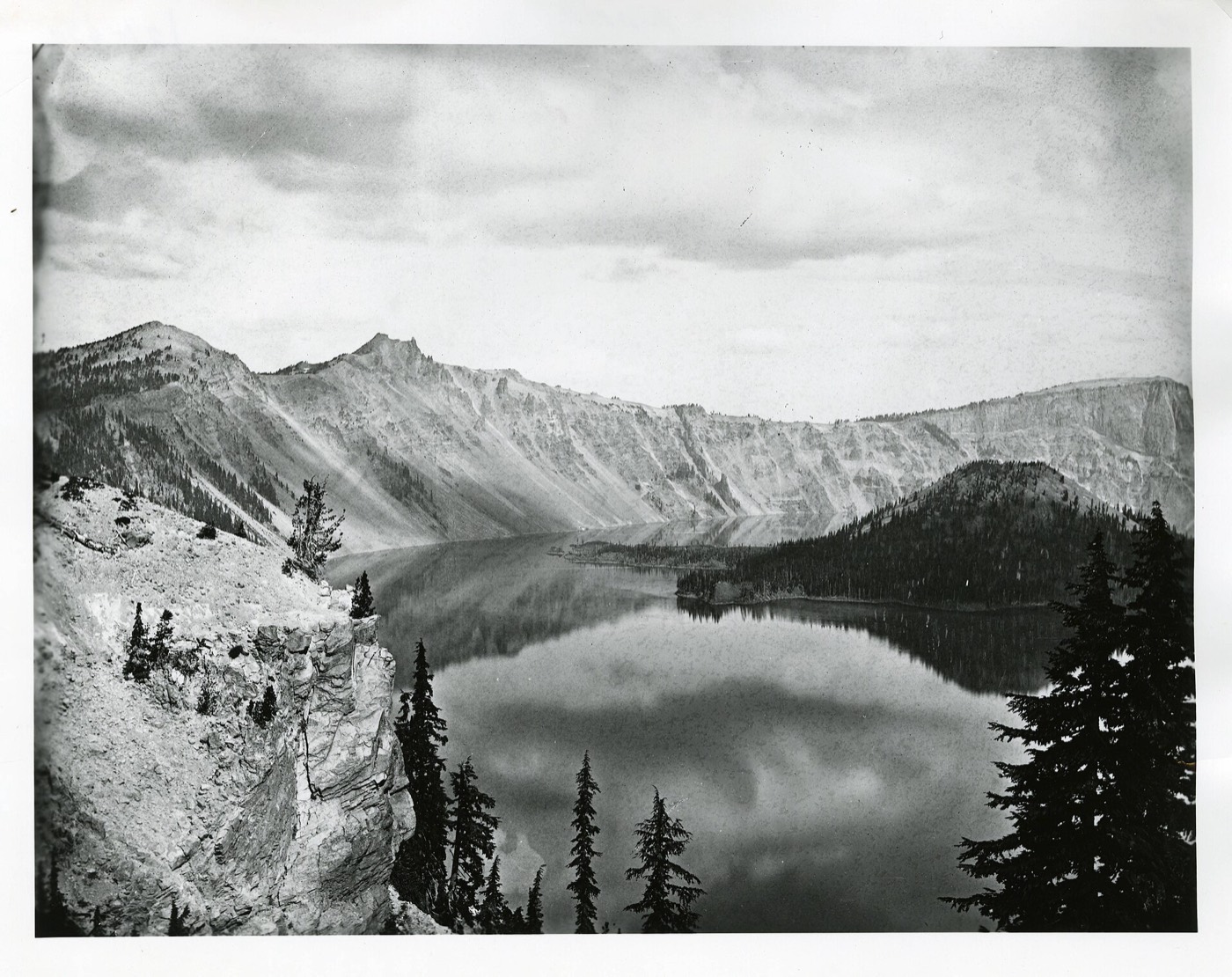An 1874 survey photograph of Crater Lake from the rim, the still water held inside the volcanic caldera with Wizard Island rising near the far shore.